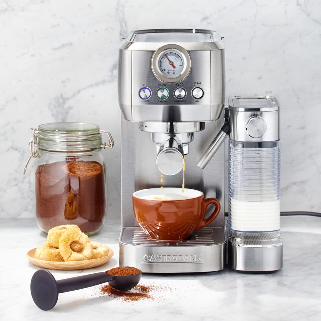 Edelstahl-Espressomaschine mit Tasse Kaffee, Milchaufschäumer und Glas mit gemahlenem Kaffee.