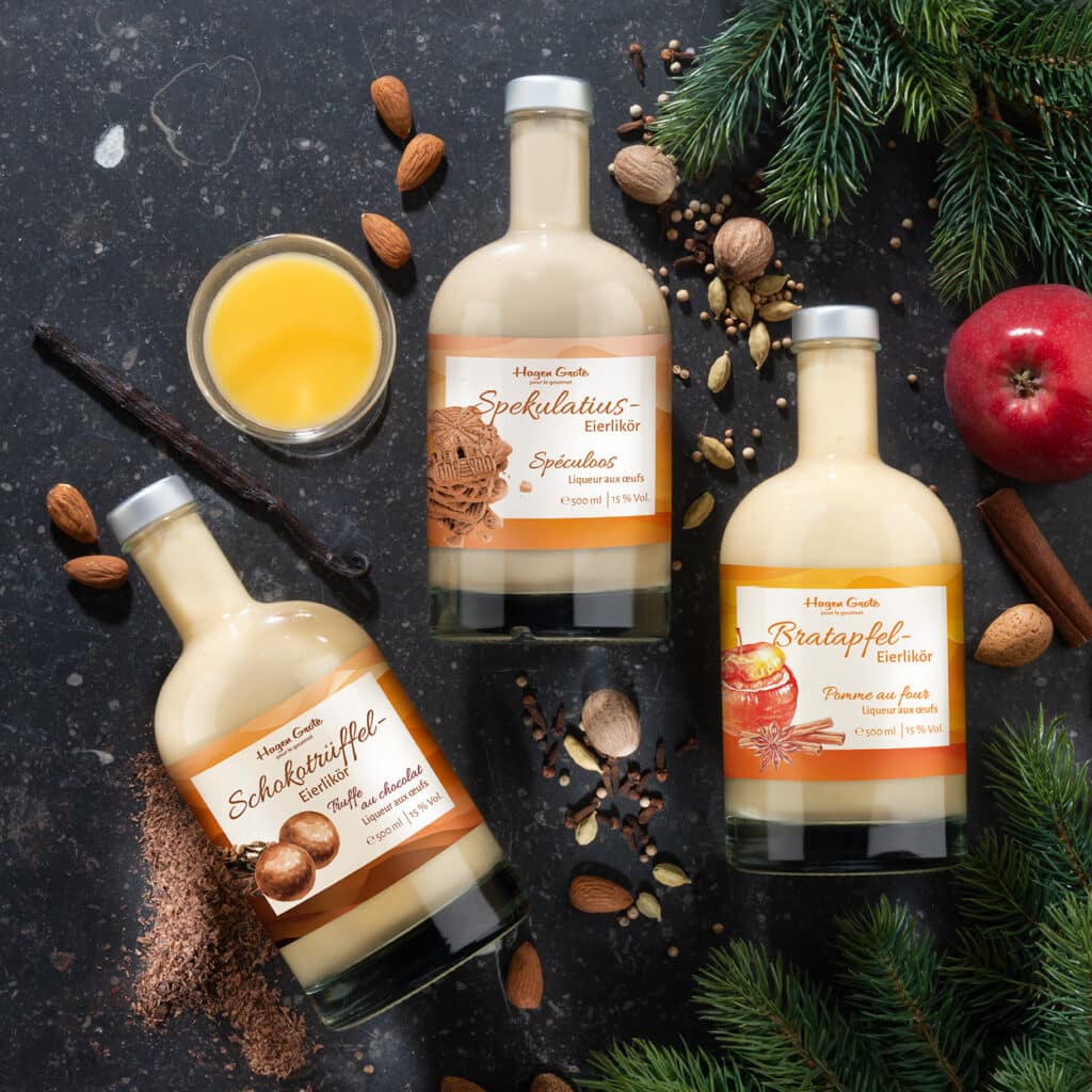 Drei Flaschen Eierlikör in Sorten Spekulatius, Schokotrüffel und Bratapfel mit Nüssen und Gewürzen arrangiert.