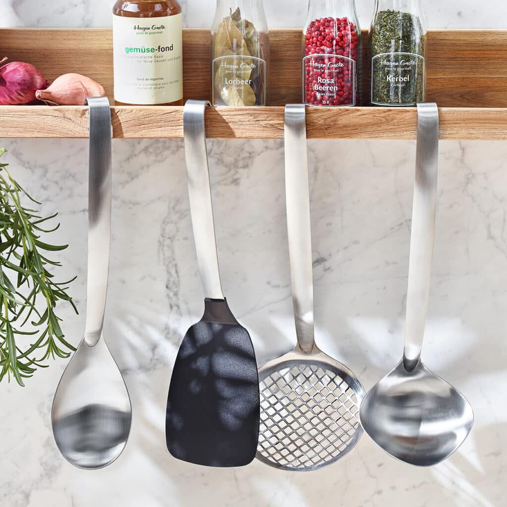 Vier Küchenhelfer aus Edelstahl am Wandregal mit Gewürzflaschen: Löffel, Schöpflöffel, Pfannenwender und Schaumlöffel.