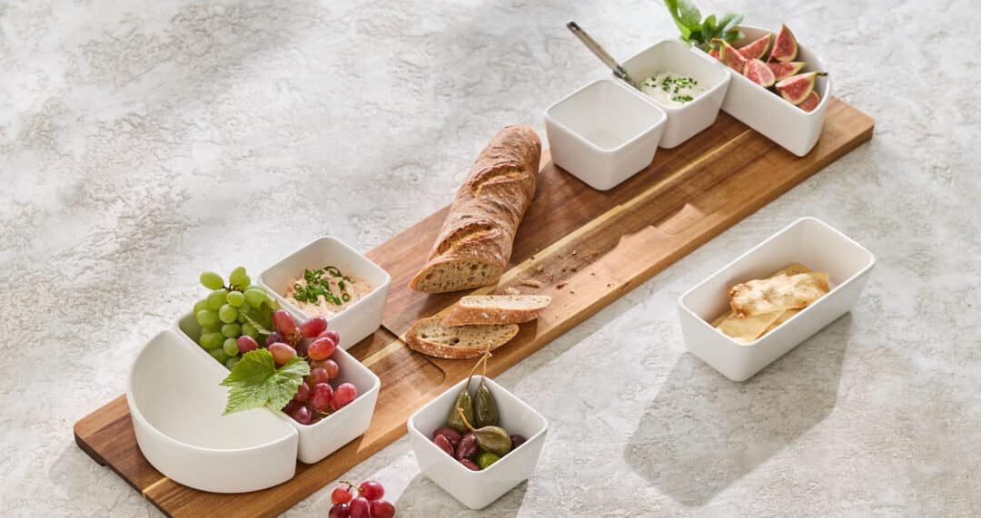 Holzbrett mit Baguette und Sch&auml;lchen, dazu Trauben, Oliven und kleine Dips in wei&szlig;en Schalen