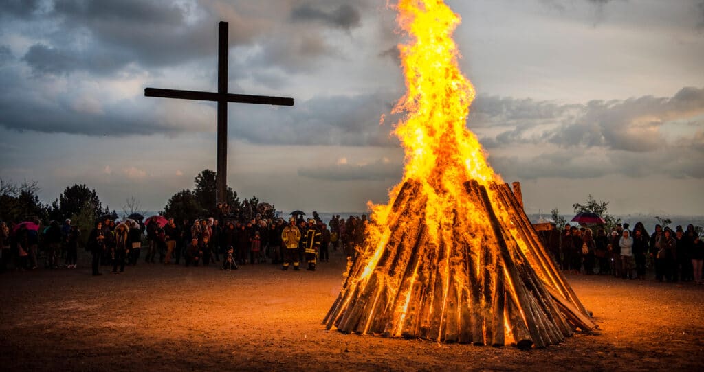 Gro&szlig;es brennendes Holzfeuer im Freien, Menschen stehen im Kreis darum, im Hintergrund ein Holzkreuz